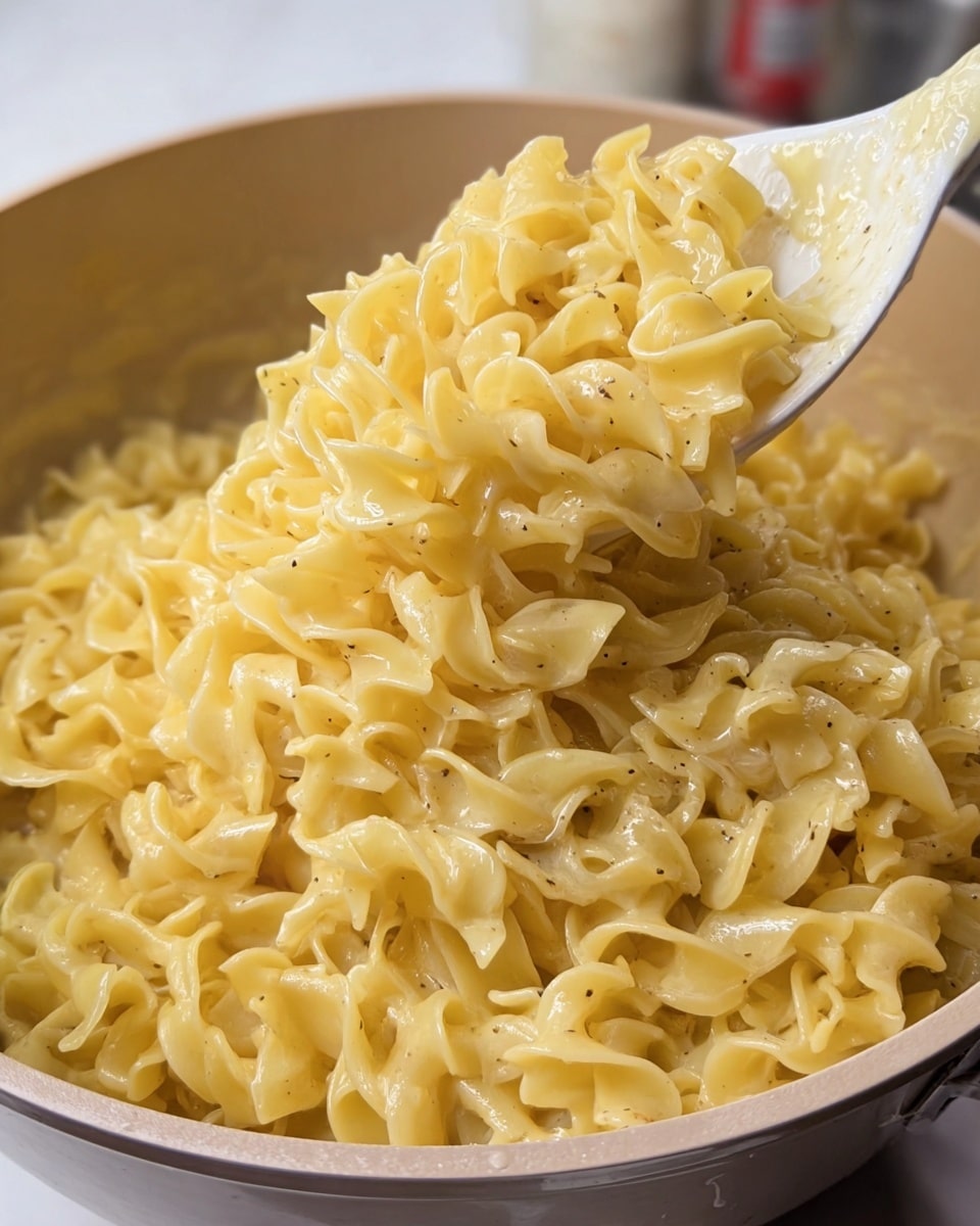 A close-up image of a pot filled with creamy cooked egg noodles that are light yellow and glossy, coated with a smooth sauce with small seasoning specks. A white spoon lifts a large scoop of the noodles, showing their twisted and curled texture with soft edges. The background includes a glimpse of a blurred container on top of a white marbled texture. The pot is light-colored and round, giving a warm home-cooked feeling. photo taken with an iphone --ar 4:5 --v 7