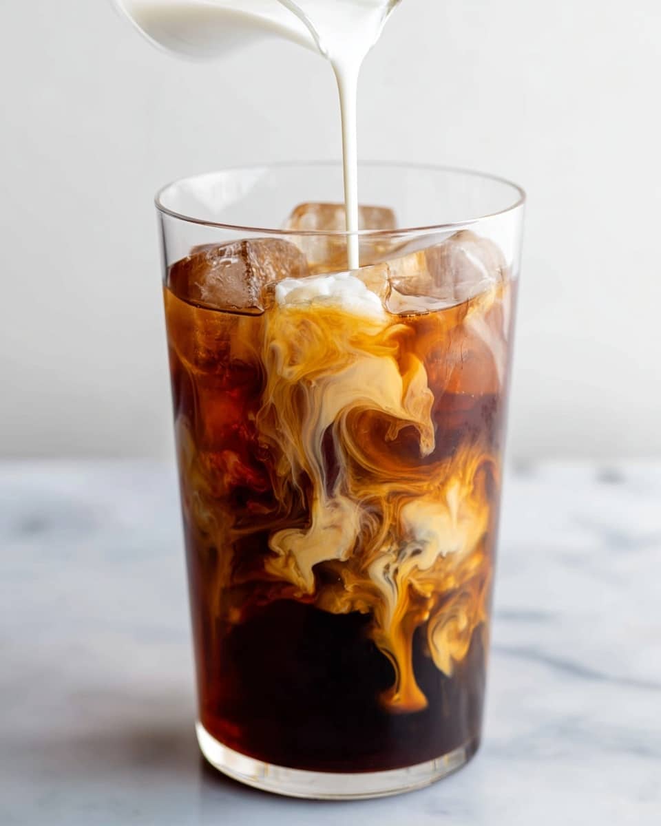 A tall clear glass filled with dark brown iced coffee and several clear ice cubes floating near the top layer. Light cream is being poured into the glass from above, creating white swirling patterns that mix with the dark coffee in the middle of the glass. The drink layers show a blend of dark and light browns where the cream meets the coffee, with intricate swirling shapes in the liquid. The glass sits on a white marbled surface. Photo taken with an iphone --ar 4:5 --v 7