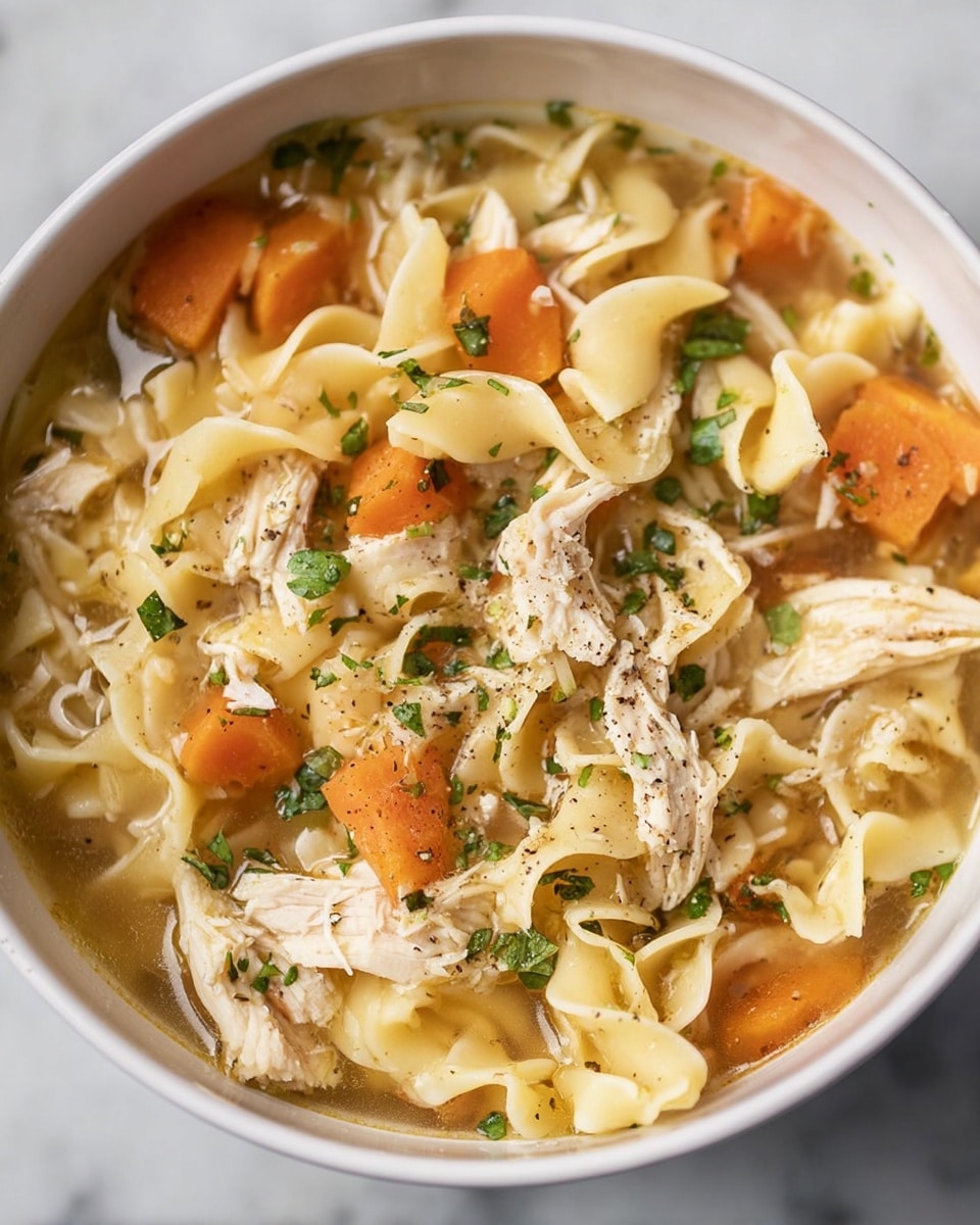 A close-up view of a white bowl filled with chicken noodle soup displaying multiple layers: at the bottom is a light golden broth, on top are chunky pieces of cooked orange carrots and shredded white chicken meat, scattered thick wide egg noodles with a pale yellow color curve softly around the bowl, and the dish is garnished with small green herb leaves with a light sprinkle of black pepper on top. The bowl sits on a white marbled textured surface. Photo taken with an iphone --ar 4:5 --v 7
