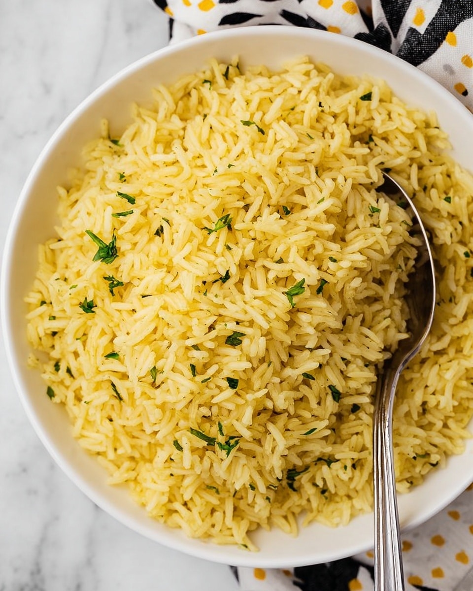 The image shows a close-up of cooked yellow rice with small green herb pieces mixed in, filling a white bowl. The rice grains look separate and fluffy, and a silver spoon partly rests in the bowl on the right side. The bowl is placed on a surface with a white marbled texture, and part of a cloth with a black and white pattern and yellow dots is visible near the top edge. photo taken with an iphone --ar 4:5 --v 7