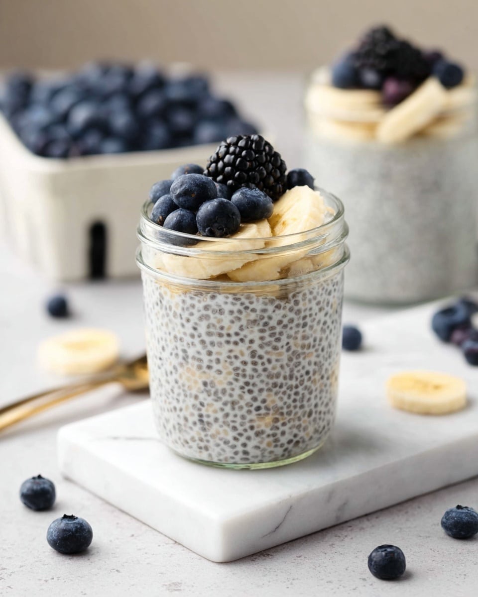 A small clear glass jar filled with two layers of creamy white chia pudding speckled with tiny dark chia seeds, topped with slices of pale yellow banana, deep blue blueberries, and a single shiny black blackberry. The jar sits on a square white marble tile surrounded by scattered blueberries and banana slices on a white marbled surface. In the background, a filled white container of blueberries is slightly out of focus, and another jar of chia pudding topped with a blackberry is visible. photo taken with an iphone --ar 4:5 --v 7