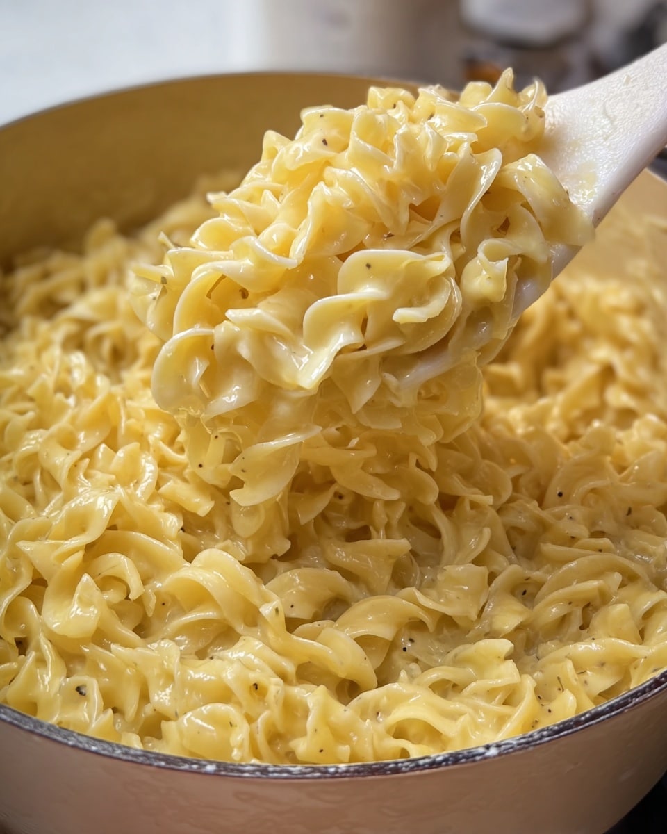 The image shows a close-up of creamy yellow pasta with a glossy texture, looking soft and coated in a thick sauce. The pasta is egg noodle shaped, with twisted, slightly ruffled edges, piled inside a white bowl. A white spoon with a wooden handle lifts a generous amount of the pasta, making the sauce shine as it catches light. The background features a smooth white marbled texture, giving a clean and simple look. photo taken with an iphone --ar 4:5 --v 7