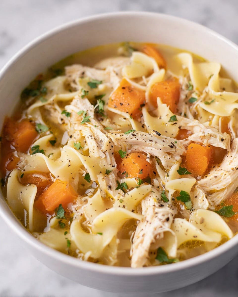 A white bowl filled with a close-up view of chicken noodle soup. The soup has multiple visible layers: chunks of light beige shredded chicken, thick pale yellow egg noodles with a slightly wavy texture, bright orange carrot slices, and small green herb pieces sprinkled on top. The broth is clear with a golden hue, and tiny black pepper specks are visible across the surface. The bowl is set on a white marbled texture background. photo taken with an iphone --ar 4:5 --v 7