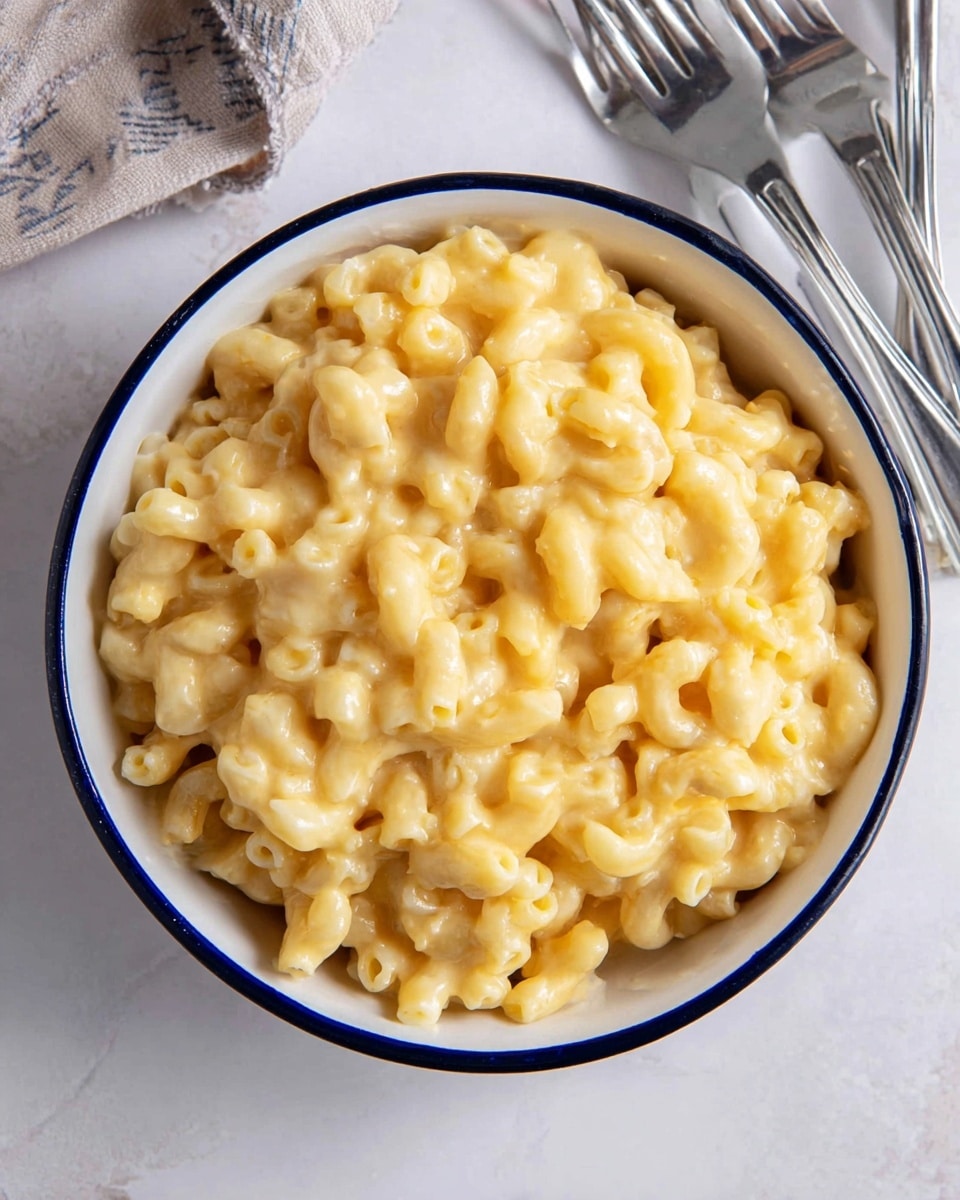 A white bowl with a blue rim filled with creamy macaroni and cheese, showing tender elbow macaroni covered evenly in a smooth, golden-yellow cheese sauce that looks slightly melted and shiny. The bowl sits on a white marbled surface, next to three silver forks arranged casually. Photo taken with an iphone --ar 4:5 --v 7