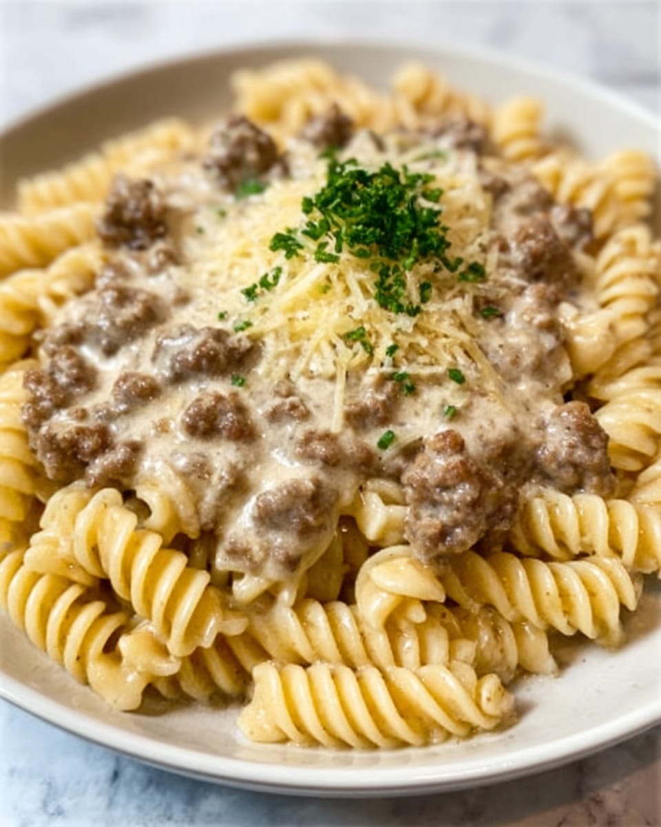 The image shows a plate of fusilli pasta with a creamy meat sauce. The pasta is light beige with a spiral texture and forms the base layer. On top, there is a thick layer of ground beef, brown and crumbly in texture, covering much of the pasta. A creamy, light yellow cheese sauce is spread over the beef, adding a smooth and rich layer. In the center, there is a small pile of finely chopped green herbs that add a fresh touch. The food is served on a white plate placed on a white marbled texture surface. Photo taken with an iphone --ar 4:5 --v 7
