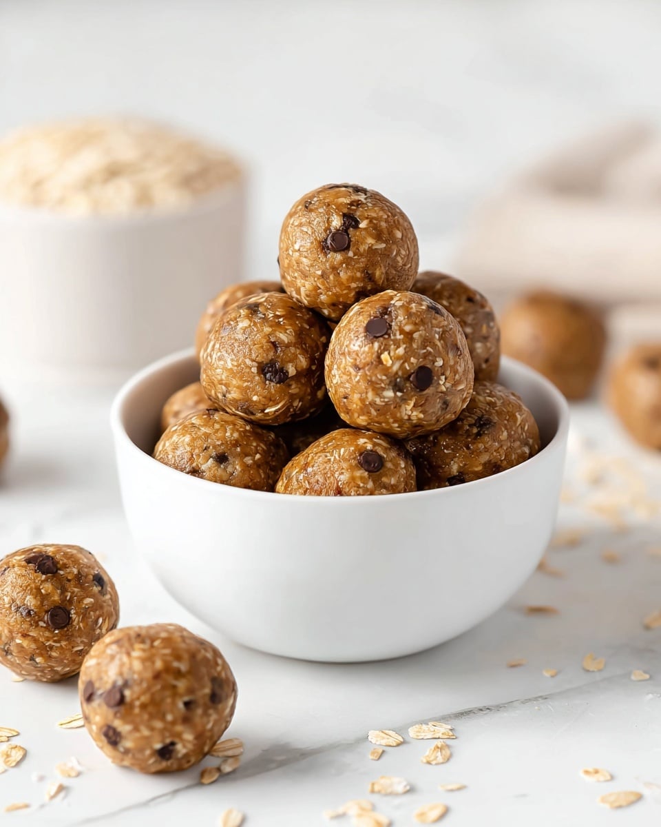 A white bowl filled with round energy balls stacked closely together, each ball showing a textured mix of brown oats and small dark chocolate chips embedded throughout. The balls have a shiny, slightly sticky surface with visible oats giving a rough texture. Around the bowl, a few more energy balls are scattered on a white marbled surface. In the blurry background, a white bowl of oats can be seen, adding context to the main dish. photo taken with an iphone --ar 4:5 --v 7