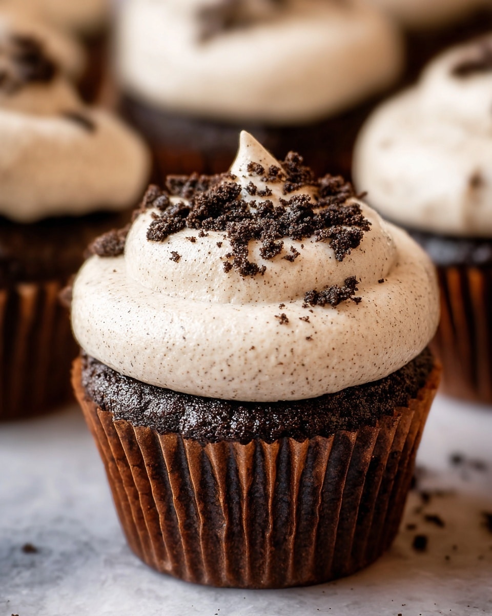 A close-up view of a chocolate cupcake with a dark brown cake base in a ridged brown paper cup. It has one thick layer of creamy, light beige frosting on top, dotted with tiny specks, and sprinkled with small dark chocolate crumbs. The cupcake is on a white marbled surface, and more cupcakes can be seen blurred in the background. photo taken with an iphone --ar 4:5 --v 7
