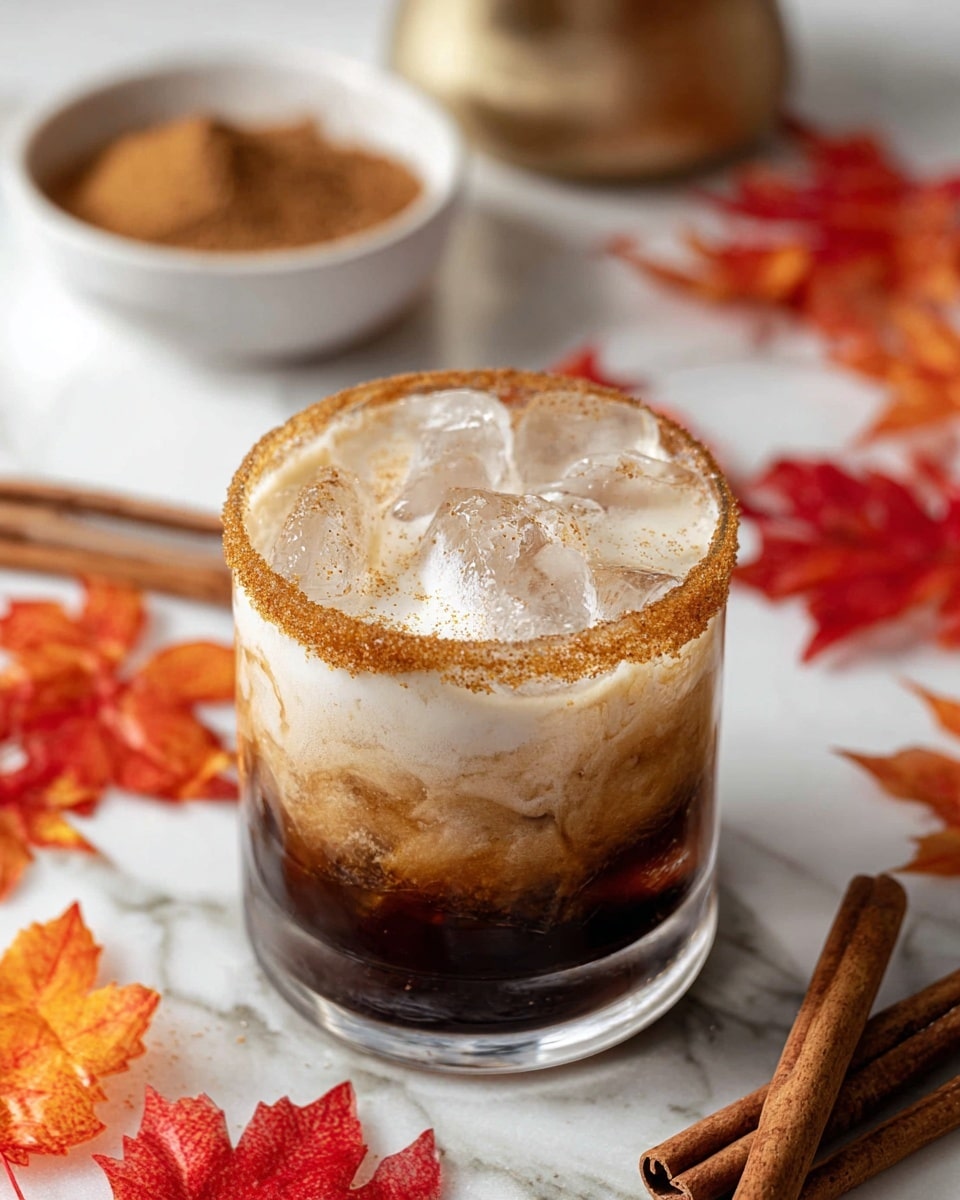 A short clear glass filled with three visible layers: a dark brown thick liquid layer at the bottom, a lighter brown frothy middle layer mixed with white foam, and a thick white foam layer on top. The rim of the glass is coated with a fine brown cinnamon powder. The glass rests on a white marbled surface with two cinnamon sticks and a red autumn leaf placed nearby. Another similar drink and some blurred bottles are visible in the background. Photo taken with an iphone --ar 4:5 --v 7