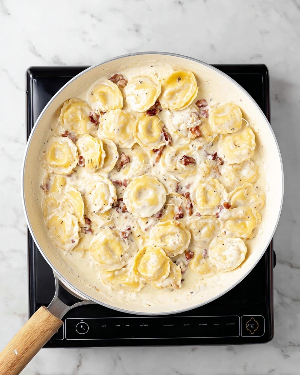 A white pan with a light wooden handle sits on a black electric stove, filled with round yellow ravioli pasta covered in a thick, creamy white sauce with small bits of reddish-brown pieces mixed in. The creamy sauce coats the pasta evenly, giving the dish a rich and smooth texture. The background is a white marbled surface. photo taken with an iphone --ar 4:5 --v 7