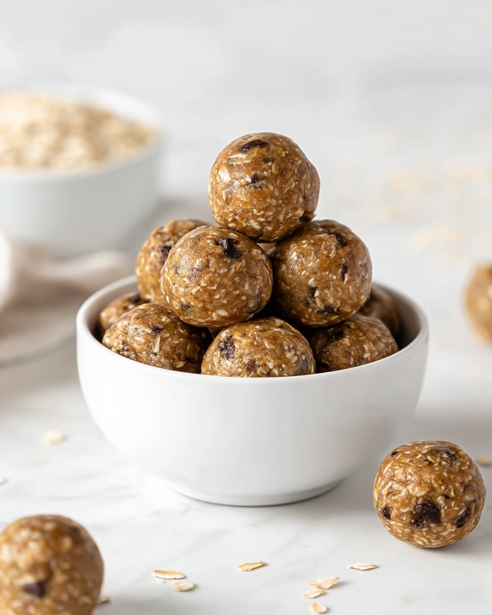 The image shows a white bowl filled with round, light brown energy balls with small dark spots, placed on a white marbled surface. Several of these energy balls are scattered around the bowl. The texture of the energy balls looks slightly rough with visible bits of oats or nuts. The lighting is soft and natural, highlighting the smooth, round shape and textured surface of each ball. photo taken with an iphone --ar 4:5 --v 7