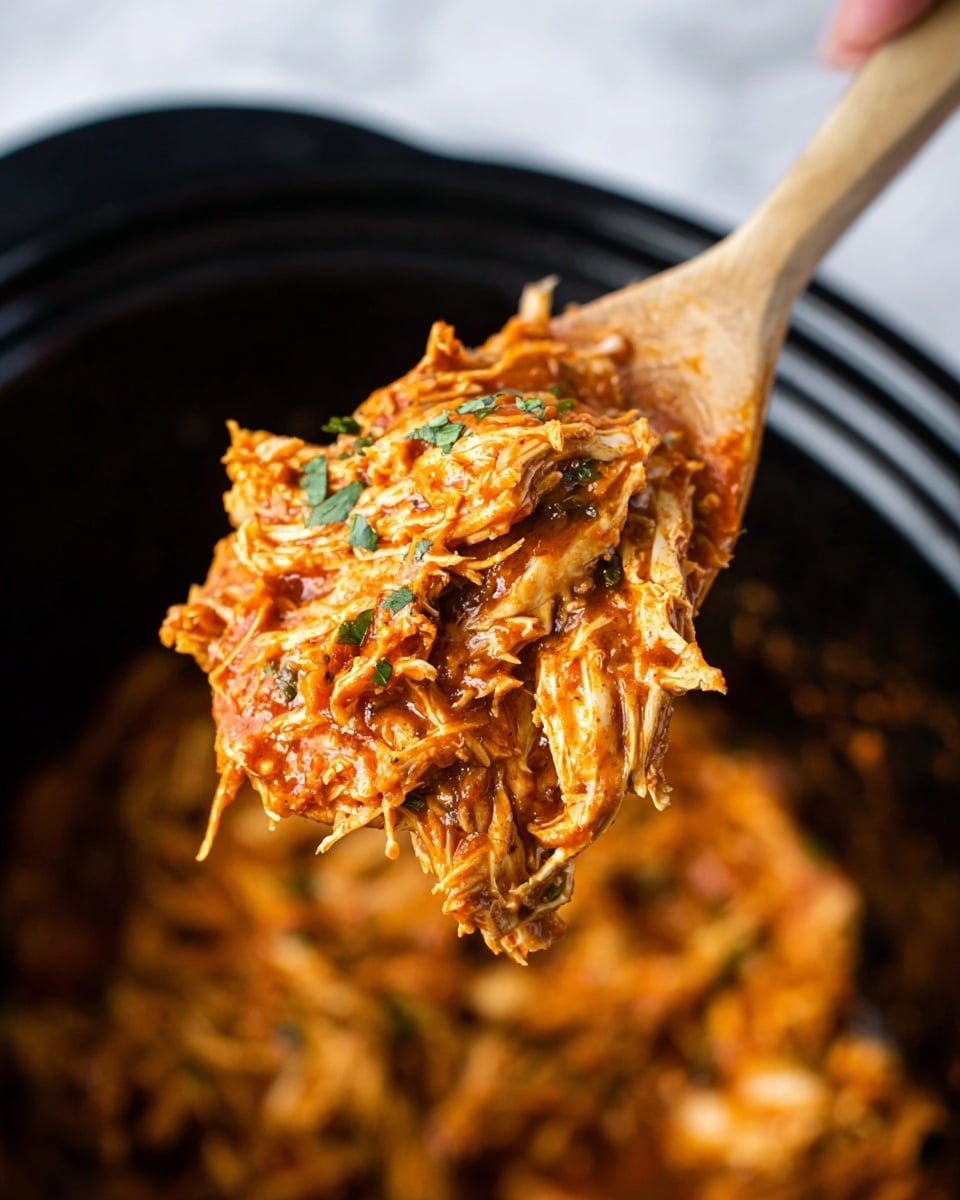 The image shows a close-up of a wooden spoon lifting shredded chicken covered in a thick, red-orange sauce with visible pieces of herbs, likely cilantro or parsley, scattered on top. The chicken strands are tender and coated evenly with the sauce, giving it a saucy and slightly glossy look. The background is a black pot filled with more of the shredded chicken mixture, creating a contrast with the rich orange color of the sauce. A woman's hand is holding the spoon at the top right corner, slightly out of focus. The scene is set on a white marbled texture surface. photo taken with an iphone --ar 4:5 --v 7