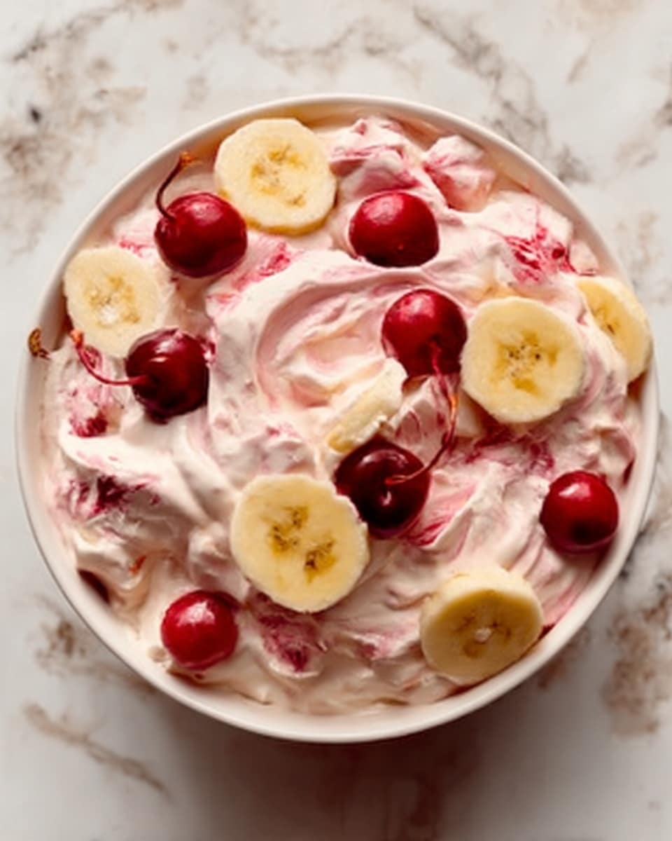 The image shows a white bowl filled with a creamy pink dessert made of small puffy pieces mixed evenly throughout. Scattered on top are bright red cherries and a few round slices of yellow banana, creating a colorful contrast. The dessert looks soft and fluffy with a smooth texture, and the bowl is placed on a white marbled surface. photo taken with an iphone --ar 4:5 --v 7