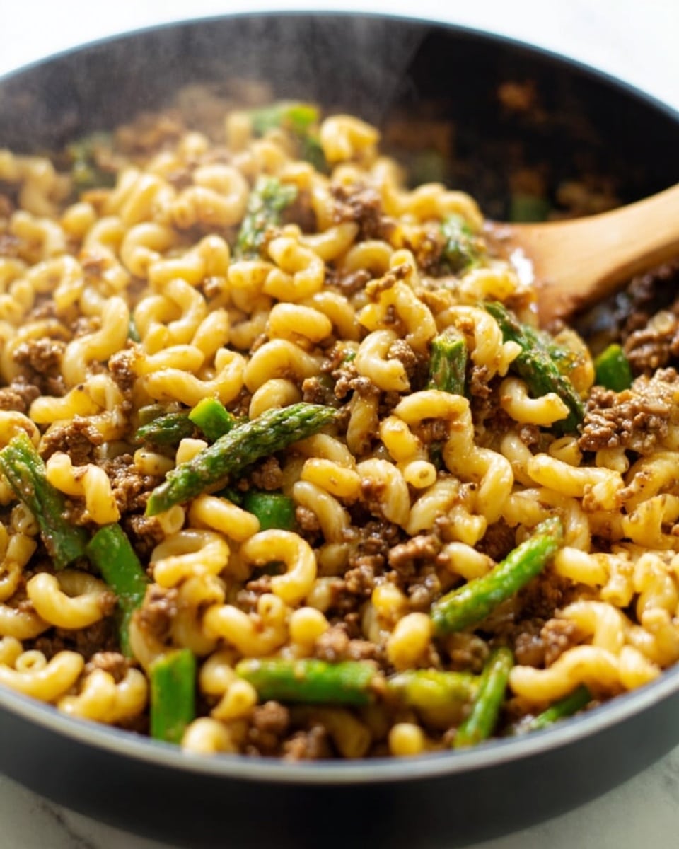 The image shows a close-up of a large pan filled with cooked elbow macaroni pasta mixed with ground meat and green asparagus pieces. The macaroni is golden with a slightly glossy texture, and the ground meat appears brown and crumbly, evenly spread throughout the pasta. The asparagus is bright green, cut into small sections, and scattered in the mix. There is steam rising from the pan, giving a warm, fresh-cooked feel. A wooden spoon is partially visible in the pan, stirring the mixture. The background is a white marbled surface. photo taken with an iphone --ar 4:5 --v 7