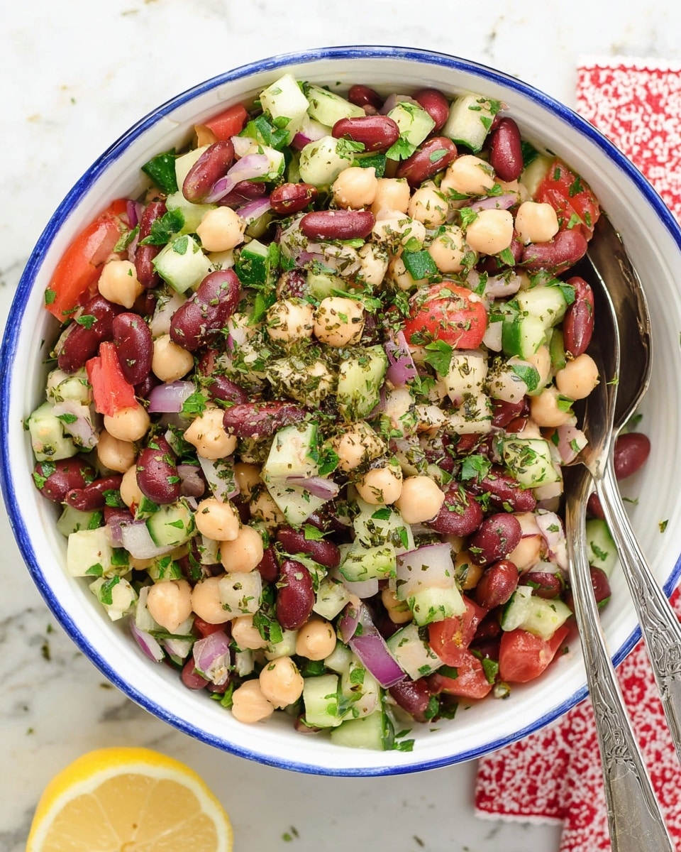 A colorful bean salad in a white bowl with a blue rim, filled with layers of red kidney beans, pale beige chickpeas, and white beans mixed with chopped green cucumber, red tomato, and purple onion. The salad is topped with finely chopped green herbs and sprinkled with dark dried herbs. Two silver salad spoons rest on top inside the bowl. The bowl sits on a white marbled surface with a red-lined cloth beneath and a lemon slice at the bottom left corner. Photo taken with an iphone --ar 4:5 --v 7
