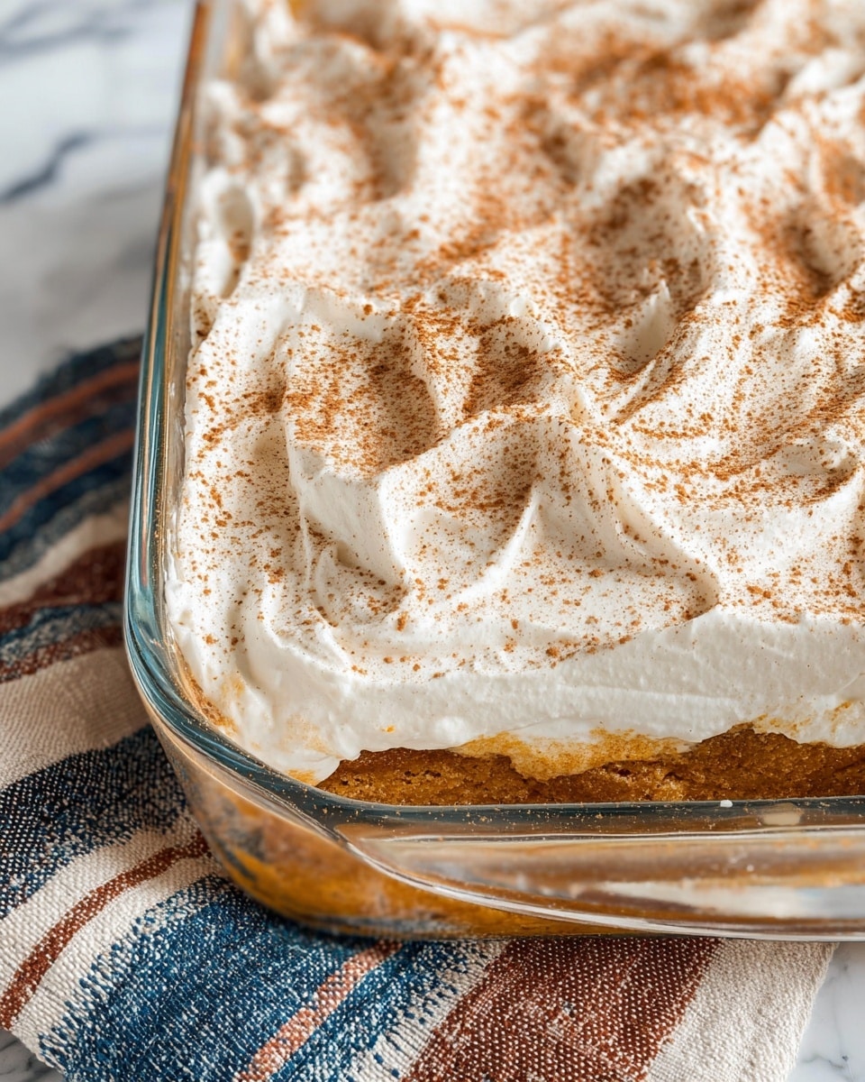 A close-up view of a dessert in a clear glass rectangular dish, showing two visible layers: a lower thick, moist, golden-brown cake layer and a top thick, fluffy white cream layer spread unevenly with texture waves. The cream layer is dusted generously with a light brown powder, likely cinnamon, adding fine specks all over. The dish sits on a white marbled surface with a textured cloth underneath featuring blue, brown, and white stripes. photo taken with an iphone --ar 4:5 --v 7