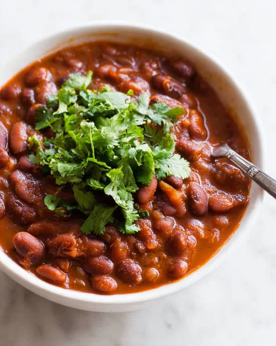 A stainless steel pot filled with a thick stew of red beans and potatoes in a rich orange-red sauce, with bright green chopped herbs sprinkled on top. The stew has visible soft potato pieces and beans throughout, creating a hearty texture. A wooden spoon with a smooth texture rests inside the pot, partly submerged in the stew, ready for stirring. The pot sits on a natural burlap cloth over a white marbled surface, and a small amount of chopped herbs lies beside the pot, adding a fresh touch. Photo taken with an iphone --ar 4:5 --v 7