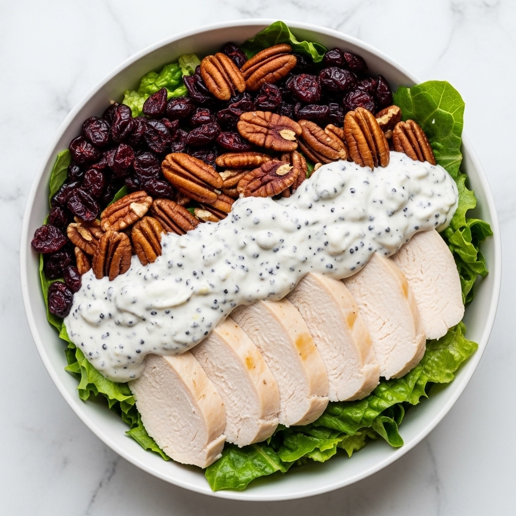 The image shows a close-up of a salad on a white plate with a white marbled background. The salad has large green leafy lettuce as the base layer spread evenly. On top of the lettuce, there is a layer of cooked chicken pieces that are creamy white with black poppy seeds sprinkled all over, giving a textured look. Scattered around the chicken are dark red dried cranberries and medium brown pecans, adding color and crunch to the dish. The overall look is fresh and rich, with a mix of creamy, crunchy, and chewy textures. Photo taken with an iphone --ar 4:5 --v 7