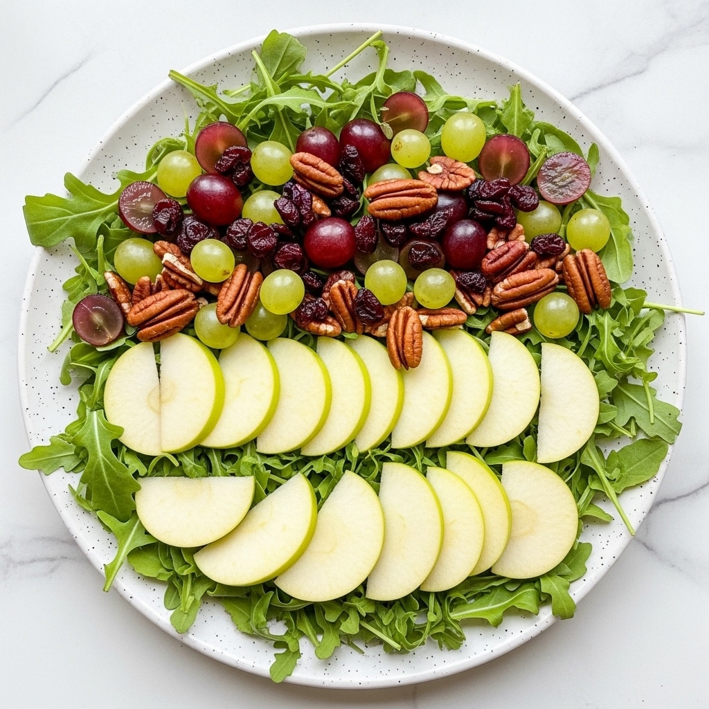 A white speckled plate holds a salad with three main layers: the base is dark green arugula leaves spread across the whole plate, the middle layer has alternating light yellow-green and pale cream apple slices arranged in a circle, and the top layer includes a scattering of whole red grapes, green grapes, dried deep red cranberries, and medium brown pecan halves and pieces, giving the dish a colorful and textured look against a white marbled surface background. photo taken with an iphone --ar 4:5 --v 7