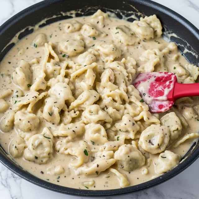 A close-up view of a black skillet filled with creamy chicken and mushroom sauce, showing tender white chicken pieces mixed with dark mushrooms in a thick yellow-orange creamy sauce, lightly sprinkled with chopped green herbs and red spices on top. The skillet handle extends out, resting on a green cloth on a white marbled surface with blurred green herbs in the background. Photo taken with an iphone --ar 4:5 --v 7