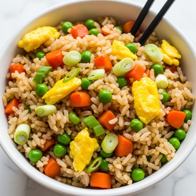 A close-up view of a bowl filled with fried rice showing many mixed colors and textures. The rice grains are light brown, mixed with small orange carrot cubes, bright green peas, chopped light green onions, and bigger pieces of yellow scrambled egg scattered all over. There are two black chopsticks standing in the bowl, partially visible. The white bowl sits on a white marbled surface, emphasizing the colorful mix inside. photo taken with an iphone --ar 4:5 --v 7