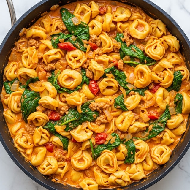 A close-up view of a large black skillet filled with creamy tortellini pasta mixed with a rich, orange-colored sauce. The dish has visible layers of green spinach leaves, browned ground meat, small pieces of red tomato, and cooked onions all blended throughout the creamy sauce. The tortellini are plump and slightly yellow, curling in various shapes across the pan. The sauce looks thick and smooth, coating all ingredients evenly. The skillet sits on a white marbled texture. Photo taken with an iphone --ar 4:5 --v 7