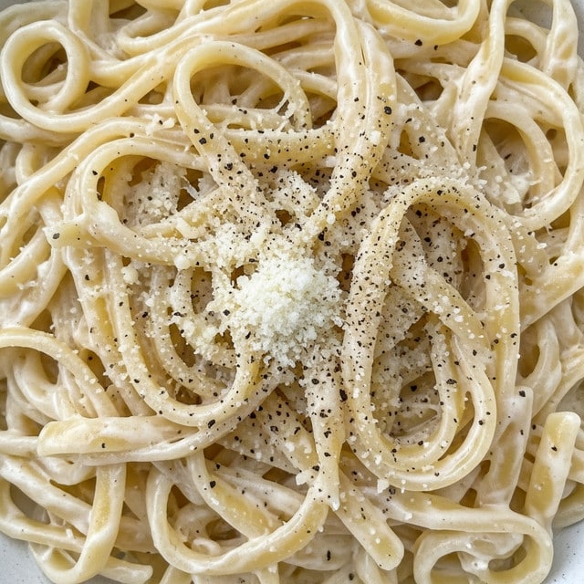 This close-up image shows creamy fettuccine pasta coated in a thick, smooth white sauce. The pasta noodles are long, flat, and slightly curled, wrapped with a rich, glossy texture. There are small bits of grated parmesan cheese sprinkled unevenly over the pasta, adding a grainy layer. Light specks of black pepper are scattered on top, creating a contrast with the white sauce. The overall look is soft, creamy, and inviting. The photo taken with an iphone --ar 4:5 --v 7