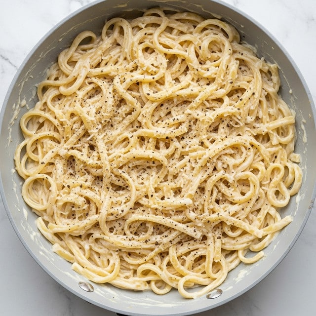 The image shows creamy fettuccine pasta in a pan with thick white sauce coating each twisted and curved strand evenly. The sauce has a smooth texture with small bits of grated cheese and is sprinkled with coarse black pepper, creating a speckled pattern on the pasta and around the edges of the pan. The pasta strands are dense and layered with glossy, rich cream, appearing soft and well cooked. The pan's inside surface is light gray, contrasting gently with the white sauce and pale yellow pasta, all set on a white marbled texture background. photo taken with an iphone --ar 4:5 --v 7