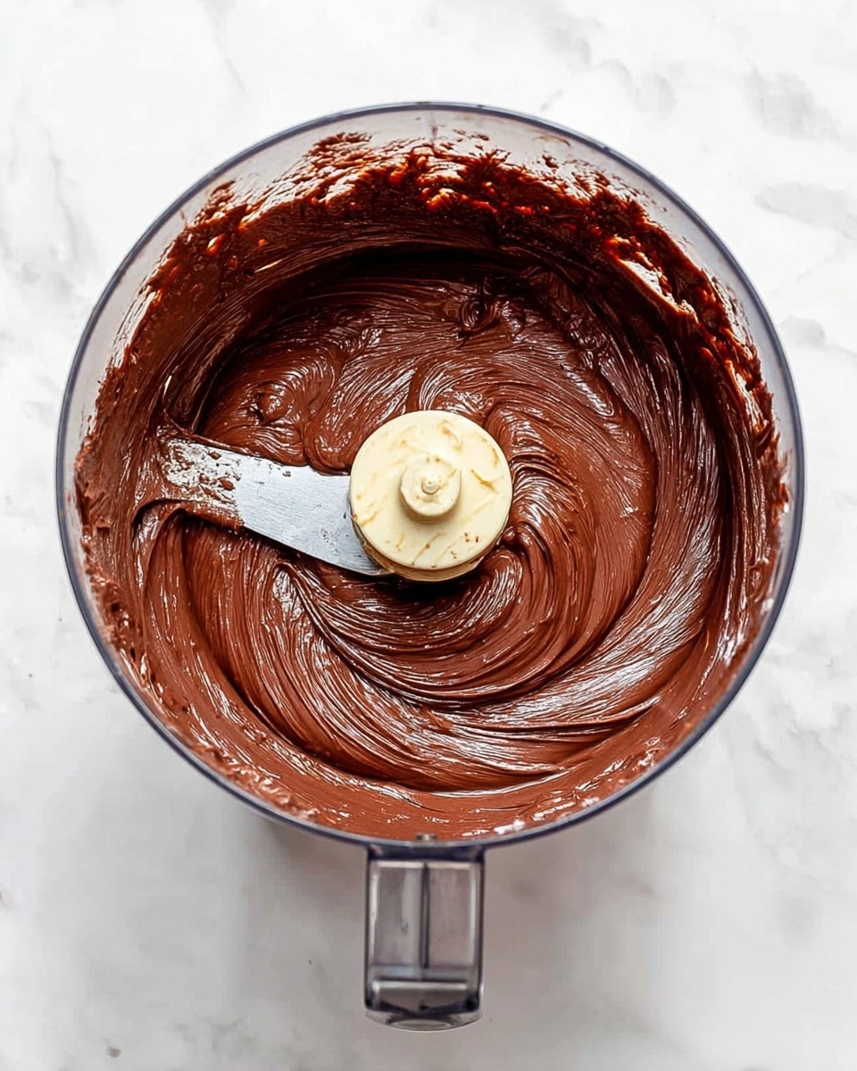 A clear food processor bowl filled with smooth, thick chocolate batter that has a shiny, creamy texture. The batter swirls around a central metal and plastic blade, with some batter clinging to the sides of the bowl, showing its rich, dark brown color. The background is a white marbled texture under the bowl. photo taken with an iphone --ar 4:5 --v 7