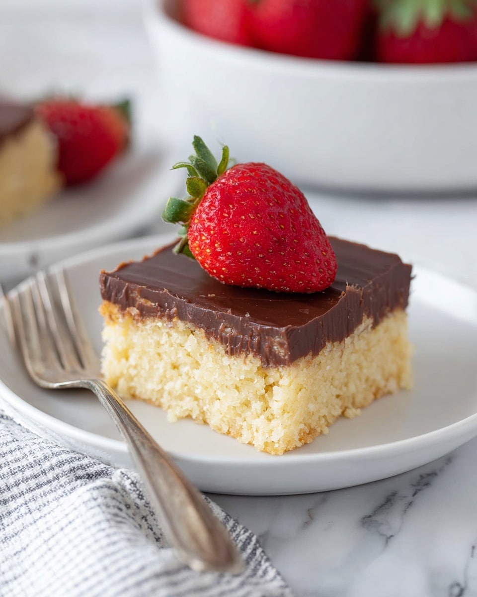 The image shows a square dessert with two layers on a white plate placed on a white marbled surface with a striped cloth underneath. The bottom layer is thick, crumbly, and light yellow, while the top layer is smooth, shiny, and dark brown chocolate. A fresh red strawberry with small seeds and green leaves sits on top of the chocolate layer. A silver fork rests beside the dessert on the plate. In the background, there is another white plate with a similar dessert and strawberry, along with a white bowl full of red strawberries, all set on the white marbled surface. Photo taken with an iphone --ar 4:5 --v 7