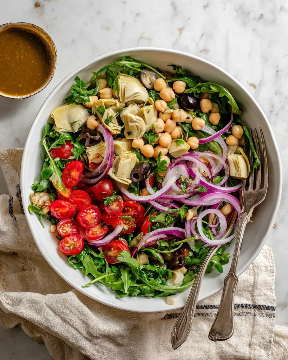 A white bowl filled with a vibrant salad showing several distinct layers: the bottom layer is fresh green arugula leaves with a leafy texture; scattered on top are chunks of light beige artichoke hearts with a soft, uneven texture; bright red cherry tomato halves add a glossy, smooth layer; beige chickpeas are sprinkled throughout, round and firm; thin slices of purple-red onion are mixed in, translucent and curved; and dark purple olives add small, shiny accents. Two silver spoons rest inside the bowl, one on the left and one on the right side. The bowl is placed on a white marbled surface with a folded beige cloth nearby and a small white bowl with light brown dressing at the bottom right. photo taken with an iphone --ar 4:5 --v 7