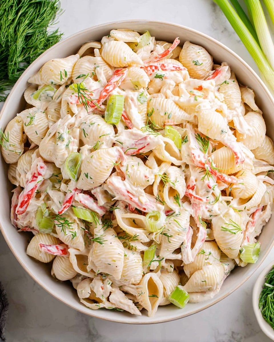 A serving of creamy pasta salad is placed on a stack of three white textured plates on a white marbled surface. The salad features pale yellow shell pasta mixed with white creamy dressing, thin slices of bright pink and white crab sticks, small pieces of green peas, and thinly sliced green onions. Small sprigs of fresh green dill are scattered on top, adding a fresh touch. In the background, fresh green onions and a halved lemon sit on the white marbled surface, along with a white cup filled with a light brown drink on a small dark coaster. Photo taken with an iphone --ar 4:5 --v 7