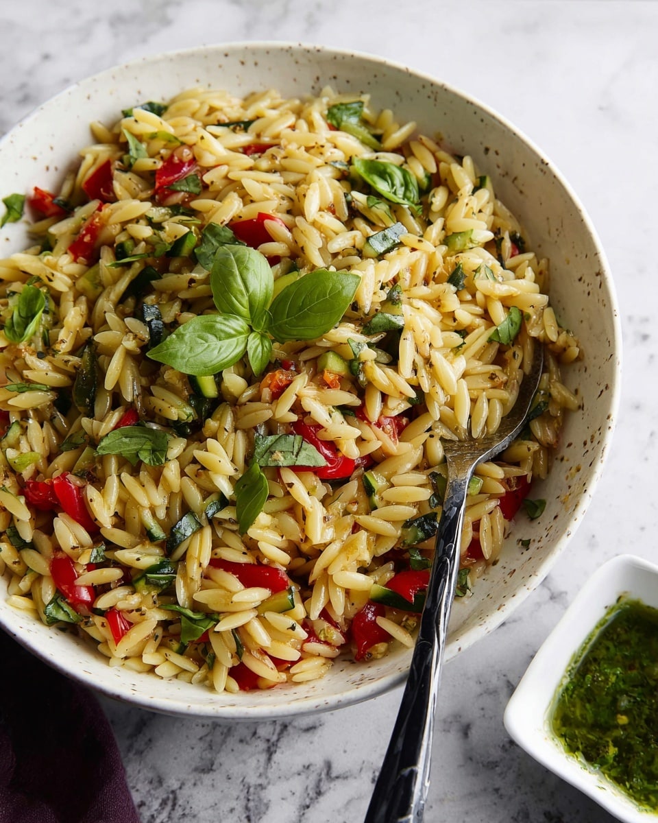 A close-up view of a spoonful of orzo pasta mixed with small pieces of green zucchini and bright red bell pepper chunks, all seasoned with black specks of herbs and spices. The orzo grains are shiny and lightly coated, appearing tender and fluffy. Green fresh basil leaves are scattered around, adding a fresh contrast. The background shows a pile of the same orzo pasta mixture on a white marbled surface. Photo taken with an iphone --ar 4:5 --v 7