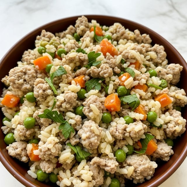 A close-up image of a dish in a brown bowl showing a mixed layer of cooked ground meat, white rice, small green peas, orange carrot cubes, and chopped green herbs. The ingredients are evenly spread and mixed, creating a textured look with soft white, green, and orange colors blending with the light brown meat. The pieces have a moist and juicy appearance, with the grains of rice and bits of vegetables visible throughout. The background is a white marbled texture. Photo taken with an iphone --ar 4:5 --v 7
