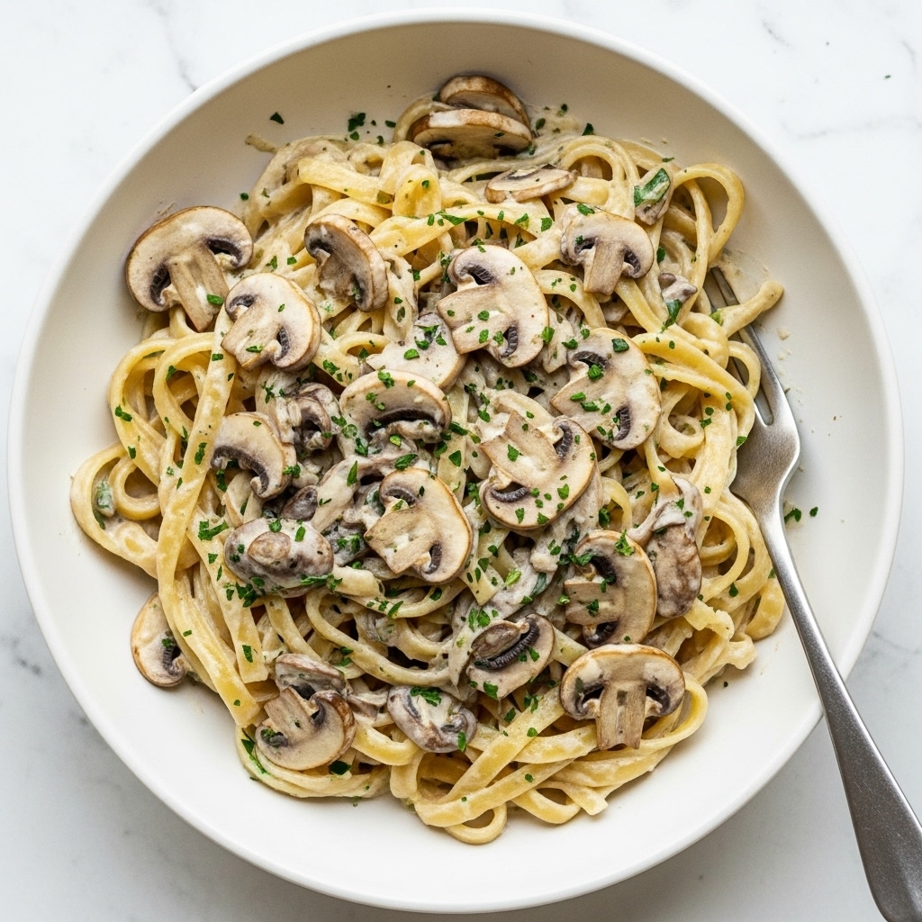 A white bowl filled with creamy mushroom fettuccine pasta is shown, with thick, flat noodles coated in a pale, smooth white sauce. There are many slices of cooked mushrooms scattered evenly on top and mixed throughout the pasta. Small green herb bits are sprinkled across the dish, adding flecks of color. A silver fork rests inside the bowl on the right side, partially under the pasta. The bowl is placed on a white marbled surface. photo taken with an iphone --ar 4:5 --v 7