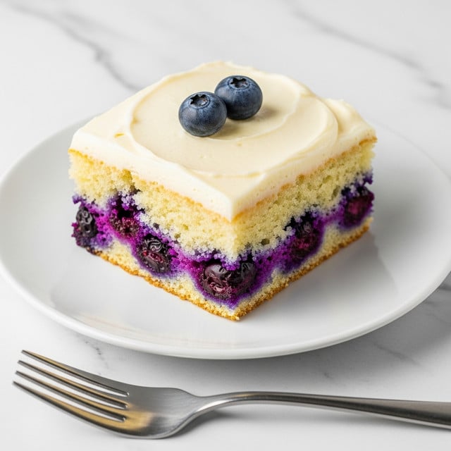 A single square piece of blueberry cake sits on a white plate over a white marbled texture. The cake has two visible layers: the bottom thick layer is light yellow with baked-in whole and broken blueberries scattered throughout, showing rich purple spots inside the soft crumb. The top layer is a smooth, creamy off-white frosting spread evenly. Two fresh whole blueberries rest on the center of the frosting. A silver fork is placed below the plate in the foreground. Photo taken with an iphone --ar 4:5 --v 7