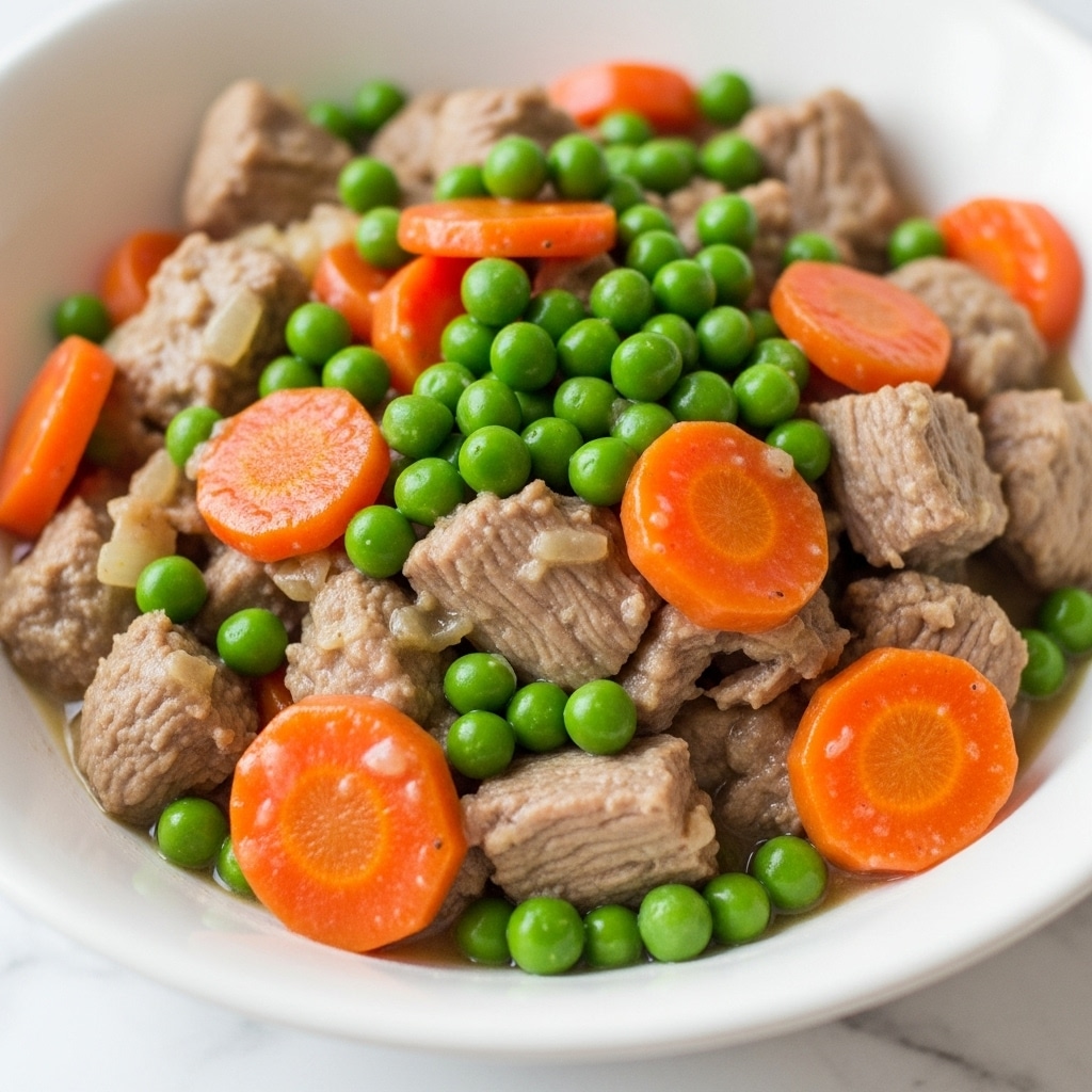 This image shows a close-up of a dish with chunks of cooked meat as the base layer that is light brown and tender in texture, topped with bright orange carrot pieces and small, round, vibrant green peas scattered evenly across the dish. The ingredients appear to be coated lightly in a pale sauce that adds a slight shine and creaminess to the overall look, all served in a white bowl set on a white marbled surface. photo taken with an iphone --ar 4:5 --v 7