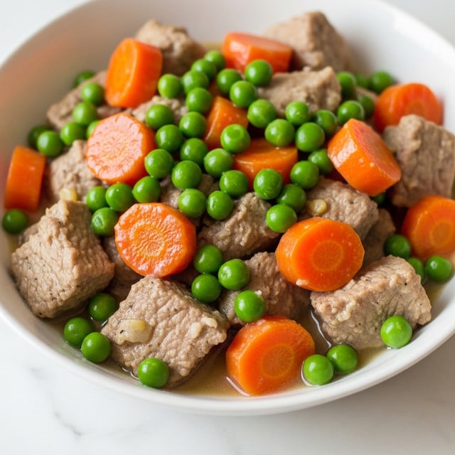 The close-up image shows a mix of cooked food layers on a white marbled surface. The first layer at the bottom has small pieces of light brown cooked meat with a rough texture. Above the meat, there are bright orange carrot chunks that look soft and slightly shiny. Bright green peas with a smooth and round shape appear scattered among the carrot pieces and meat. The overall image is colorful and the food looks moist and tender. Photo taken with an iphone --ar 4:5 --v 7