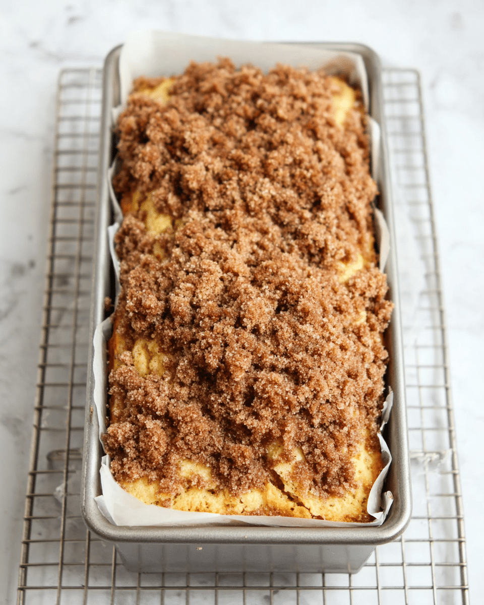 The image shows a loaf-shaped cake with two main layers visible from the top: a golden yellow cake base layer and a thick crumbly brown streusel topping. The streusel is unevenly spread, creating a rough, textured surface in various shades of brown. The cake is baked inside a silver metal pan lined with white paper and placed on a wire cooling rack. The background is a white marbled texture. photo taken with an iphone --ar 4:5 --v 7