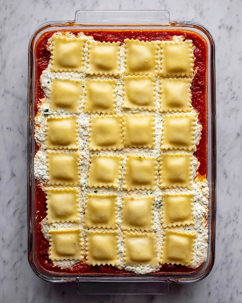 A clear rectangular glass baking dish holds a layered ravioli casserole. The bottom layer is a bright red tomato sauce, partially visible along the edges. On top of this is a creamy white cheese mixture with bits of green herbs peeking out. The top layer consists of neatly placed square ravioli pieces arranged in a grid, each pale yellow with a slightly rough texture and zigzag edges, covering the dish completely. The whole setup is on a white marbled surface. photo taken with an iphone --ar 4:5 --v 7