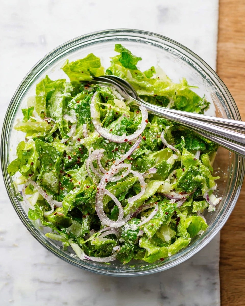 A clear glass bowl filled with a fresh salad showing layers of bright green chopped lettuce and thin, spiral slices of light purple onion scattered throughout. Small white crumbs are sprinkled all over the top, along with tiny red pepper flakes for color contrast. Two silver salad forks rest inside the bowl, one leaning on the edge, ready to serve. The bowl is placed on a white marbled texture surface, adding a clean and fresh backdrop. Photo taken with an iphone --ar 4:5 --v 7