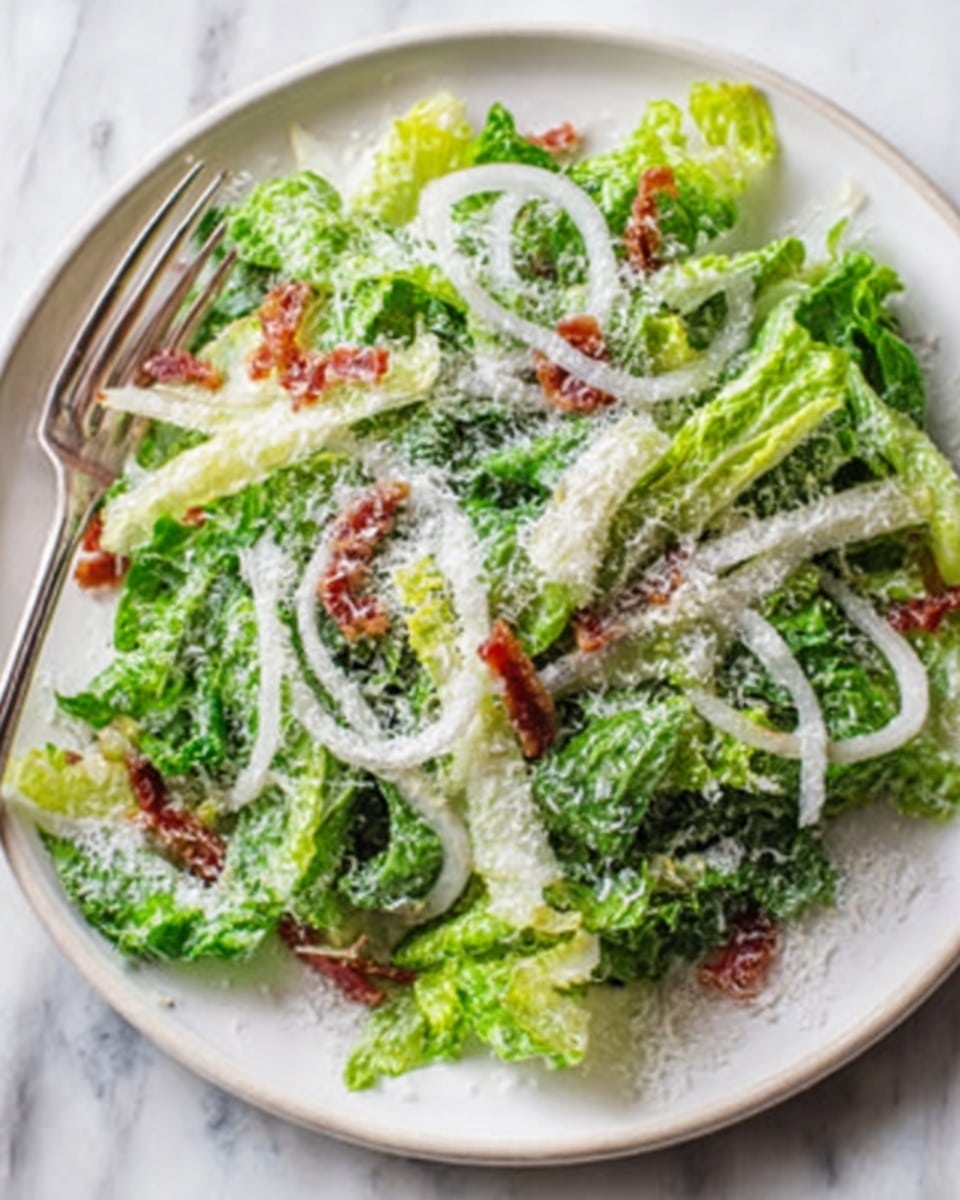A fresh salad is served on a white plate sitting on a white marbled surface, featuring several layers. The base layer is a mix of light and dark green leafy lettuce, cut into long thin strips. On top are thin white rings of raw onion scattered evenly. Small pieces of reddish-brown bacon or similar ingredient are spread across the salad, adding texture. The entire salad is finished with a generous, fine sprinkle of white grated cheese dusted all over, giving a slightly fluffy look. A silver fork is placed on the left edge of the plate, ready to eat. Photo taken with an iphone --ar 4:5 --v 7