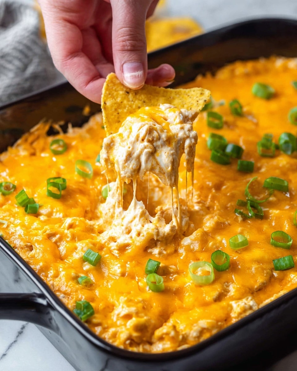 A close-up view of a black rectangular dish filled with a thick, creamy cheesy dip that is bright orange with melted cheddar cheese layered on top, mixed with shredded white cheese underneath, creating a textured surface. Scattered thinly sliced green onion rings add a pop of fresh green color across the dish. A woman's hand is holding a triangular yellow corn chip dipped into the gooey cheese, showing stretchy melted cheese strands pulling from the dip to the chip. The dish rests on a white marbled textured surface. photo taken with an iphone --ar 4:5 --v 7