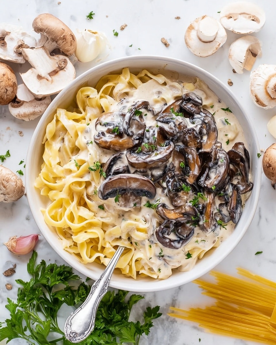 A white bowl filled with three layers: at the bottom, light golden fettuccine pasta strands arranged loosely; on top of the pasta, creamy white mushroom sauce with visible slices of light brown and dark brown mushrooms; sprinkled over the dish is a small amount of finely chopped green parsley. A silver fork rests inside the bowl, poking into the pasta. The background is a white marbled surface with scattered garlic cloves, fresh parsley, and a small bunch of uncooked fettuccine pasta. Photo taken with an iphone --ar 4:5 --v 7